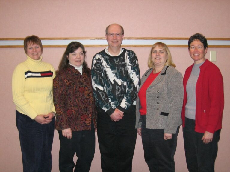 four middle-aged white females and one middle-aged male in early 2000's fashion standing in front of a wall.