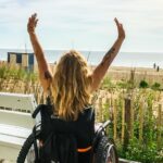 a woman in a wheelchair raises her hands to the sky. In the background is the beach.