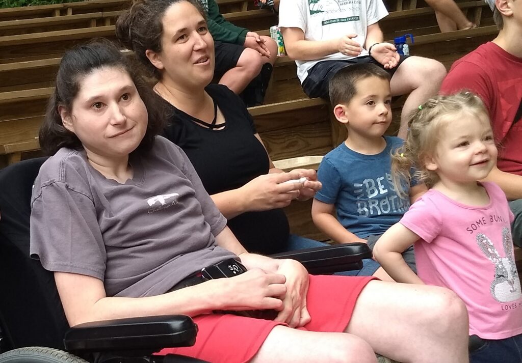 Emily in a wheelchair sitting with her sister and two niblings at the ampitheater