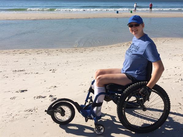 A woman in a wheelchair smiles at the camera on the beach with the ocean in the background