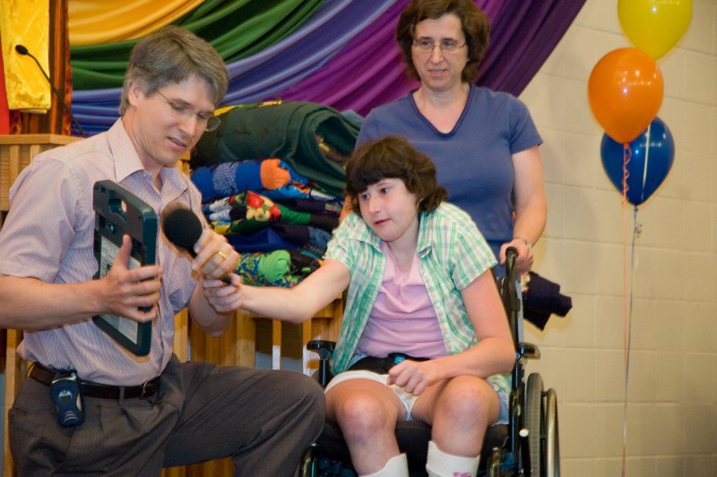 A young woman in a wheelchair speaks through an ipad while her mother and father stand next to her, with colorful fabric in the background