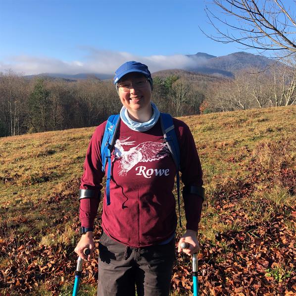 A woman in a red shirt smiles at the camera, with forearm crutches and mountains in the background