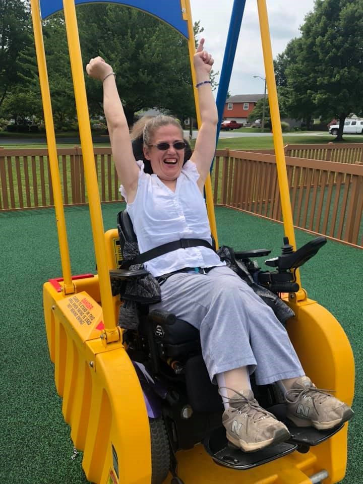 Woman swinging on an adaptive swing in her power wheelchair, smiling with her arms raised