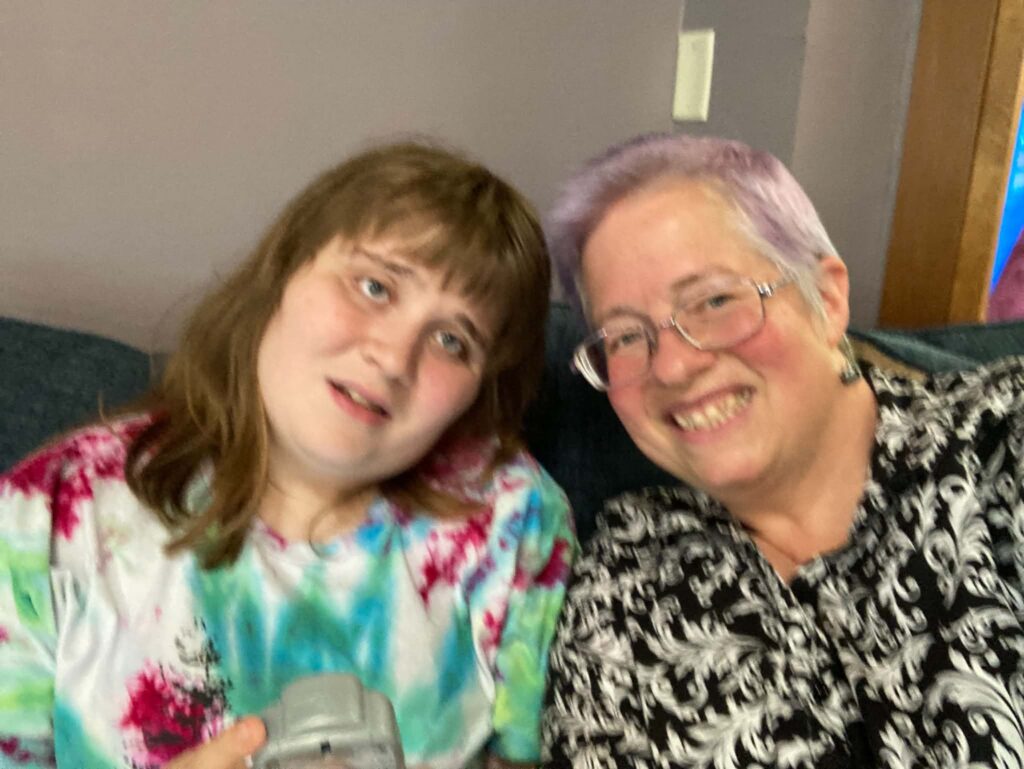 Two white women smiling at the camera, sitting on a blue couch. One of them is wearing a tie dye t-shirt and the other is wearing a black and white floral print collared shirt