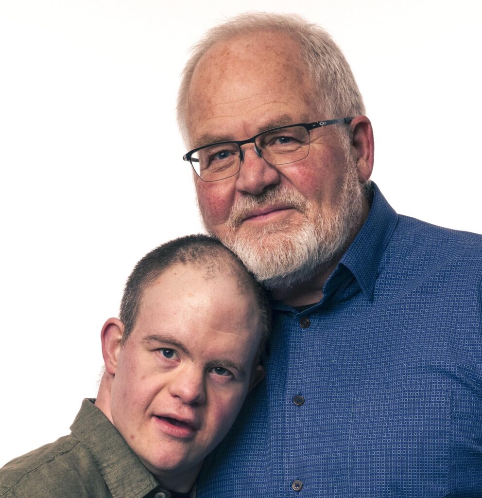 A father and son pose for the camera. The son has Down syndrome and has short brown hair and is wearing a green collared shirt. The father has short white/gray hair and glasses and is wearing a blue collared shirt.