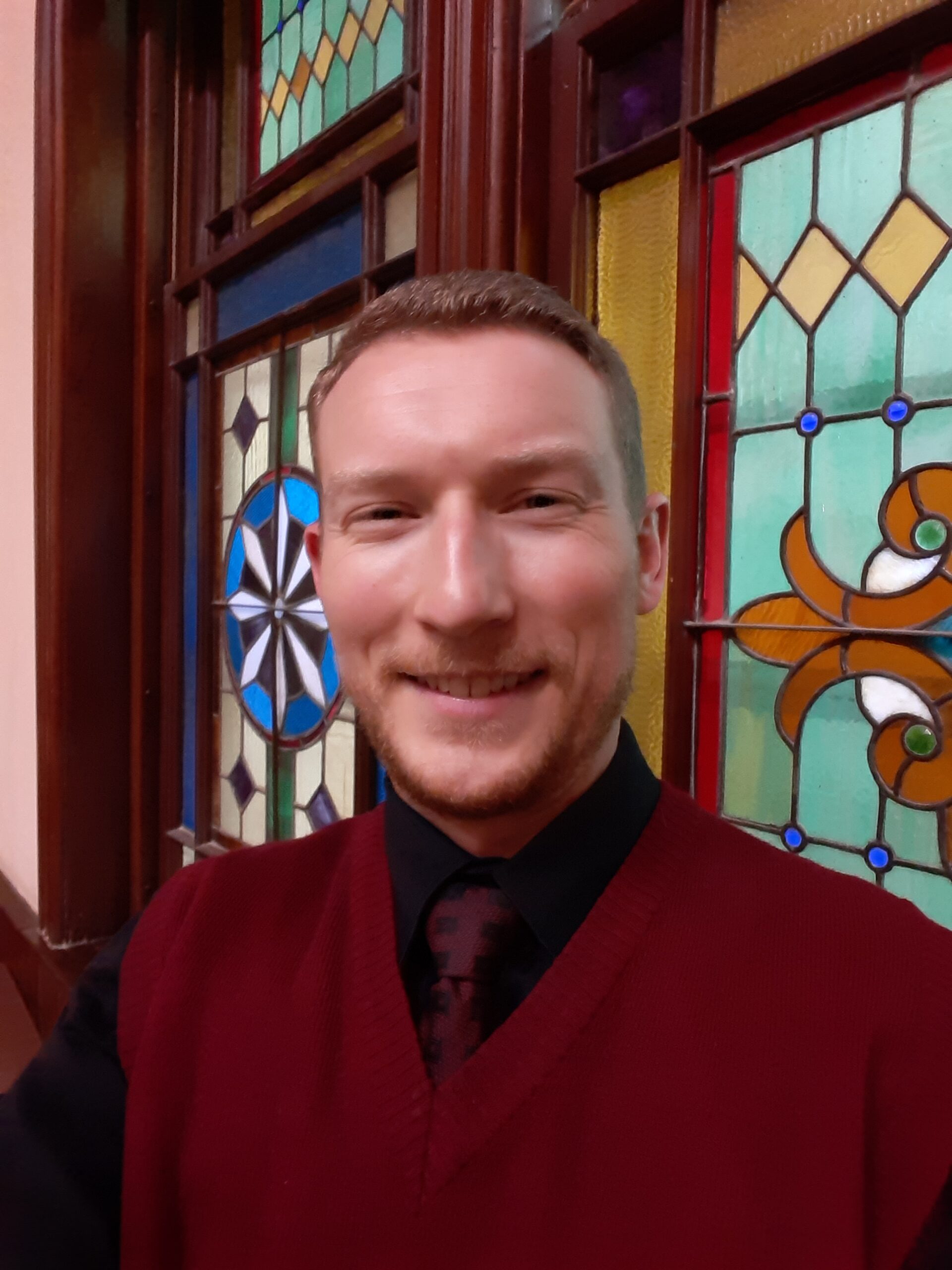 an image of a white man with short hair wearing a red sweater with stained glass windows in the background